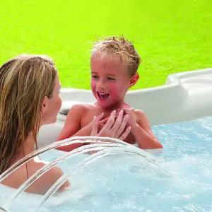 A woman and a young child smile and play together in a bubbling outdoor hot tub, with green grass visible in the background. Water arcs from small jets in the foreground.