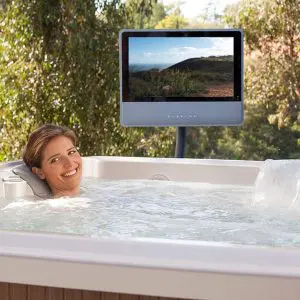 A woman relaxes in an outdoor hot tub, smiling at the camera. Behind her, a television screen displays a landscape scene with hills and greenery. Trees are visible in the background.