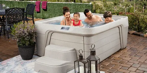 A smiling family of four relaxes together in a light-colored hot tub on a patio, surrounded by plants, a dining set, a rug, and decorative lanterns.