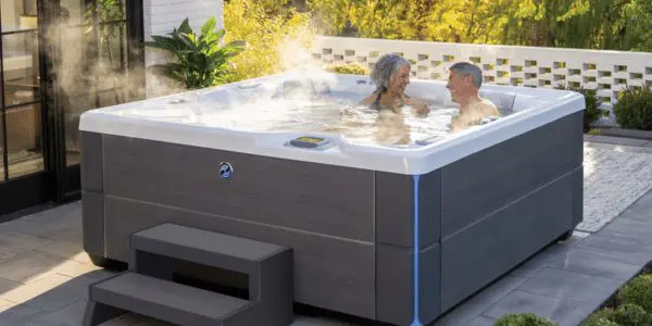 An older couple smiles and chats while relaxing in a steaming outdoor hot tub on a patio, surrounded by greenery and modern decor.