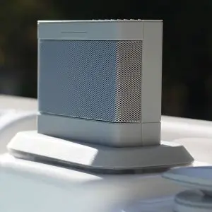 A close-up of a portable gray wireless speaker with a mesh front, placed on a smooth white surface outdoors.