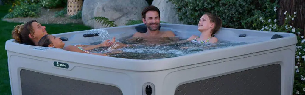 Four people relax and smile together in a square outdoor hot tub surrounded by greenery, with water bubbling gently around them.