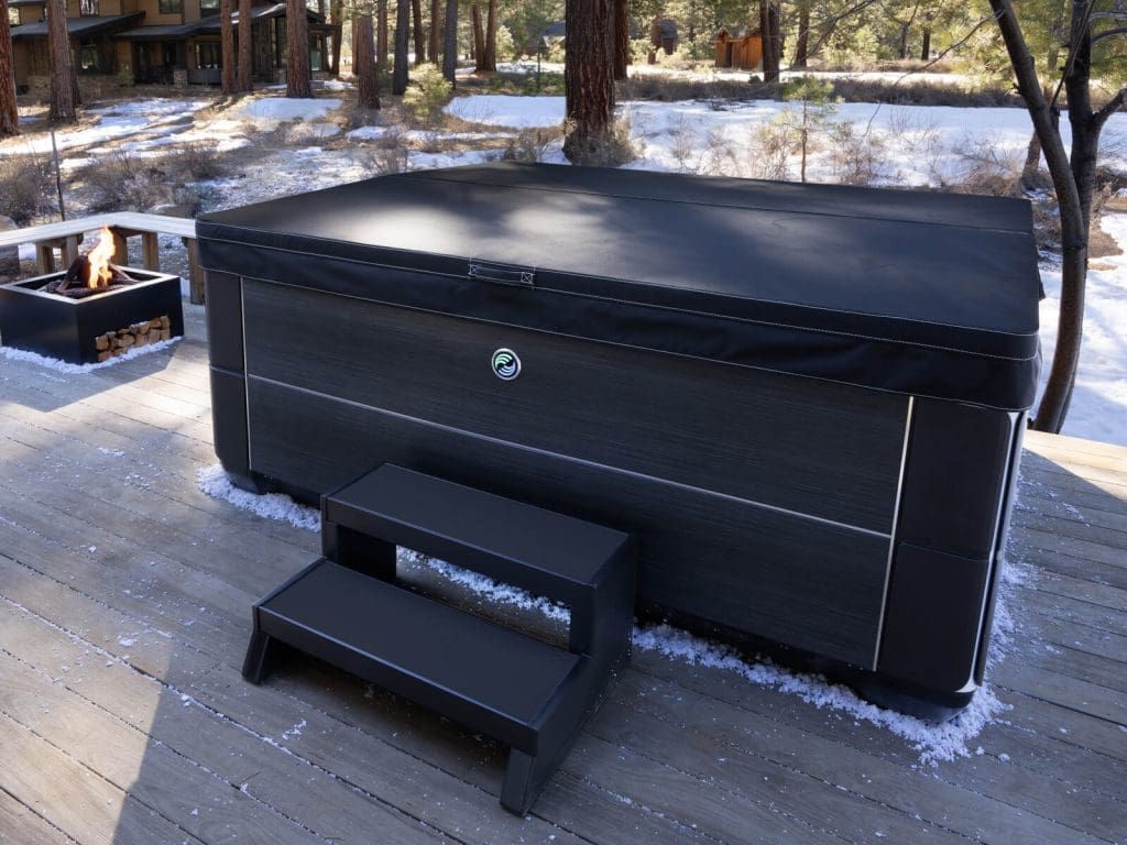 A covered hot tub with black steps sits on a wooden deck outdoors, surrounded by trees and light snow. A square fire pit with stacked wood is nearby.