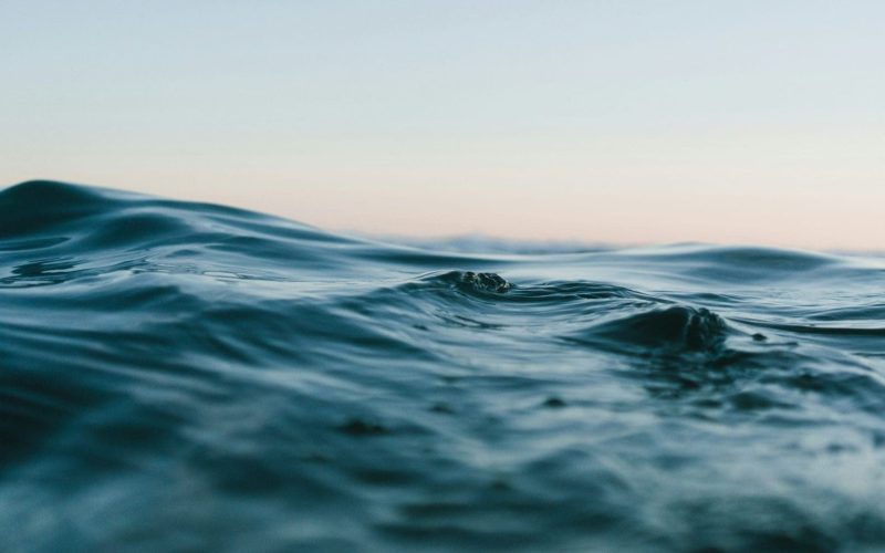 Close-up view of gentle ocean waves with a soft blue hue, under a clear sky with a faint gradient from light pink to blue at the horizon.