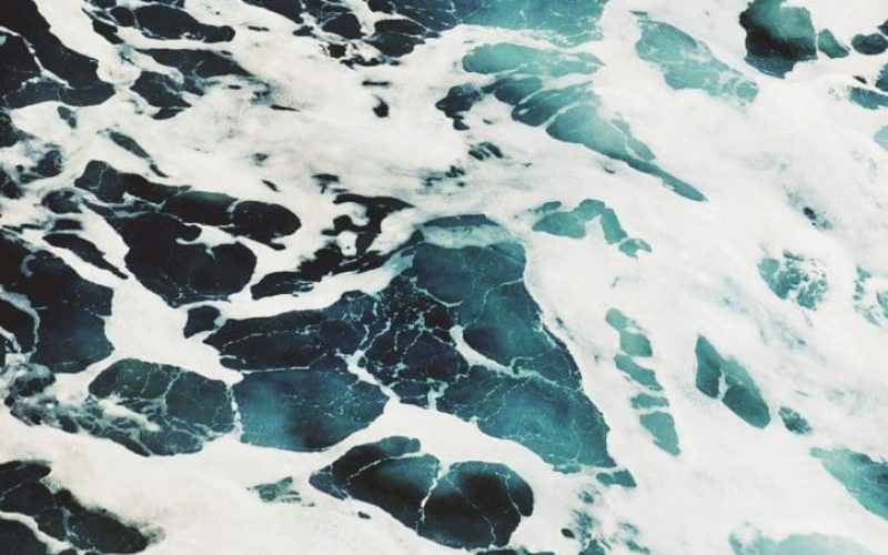 Aerial view of ocean waves with white foam contrasting against deep blue and turquoise water, creating an abstract, swirling pattern.