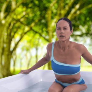 A woman in a blue one-shoulder swimsuit relaxes in a hot tub outdoors, surrounded by lush green trees and sunlight filtering through the leaves.
