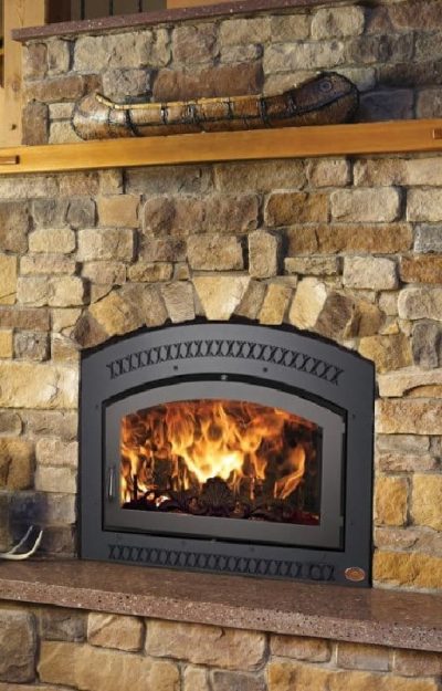 A stone fireplace with a burning fire behind a black metal grate, a brown ceramic vase on the hearth, and a wooden ledge above, set in a cozy living room with a staircase in the background.