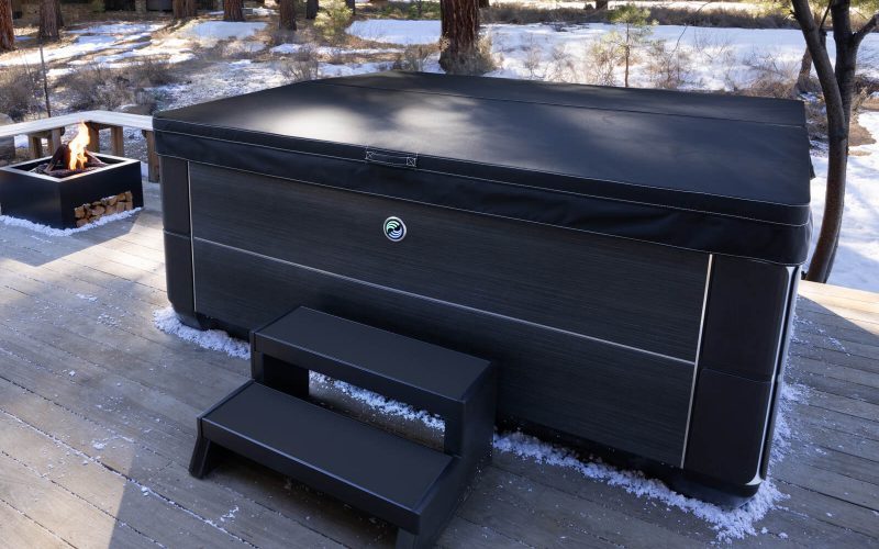 A covered hot tub with black steps sits on a wooden deck outdoors, surrounded by trees and light snow. A square fire pit with stacked wood is nearby.