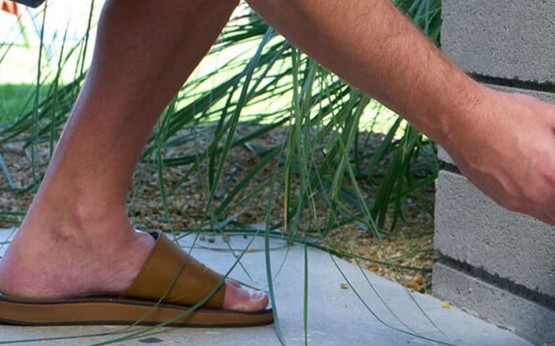 A person wearing sandals and shorts plugs a black electrical cord into an outdoor wall outlet beside a concrete block wall.