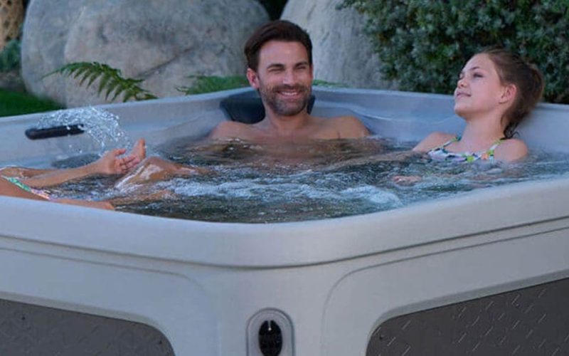 Four people relax and smile together in a square outdoor hot tub surrounded by greenery, with water bubbling gently around them.