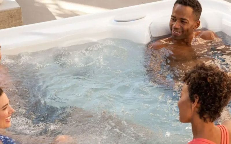 Four people relax and smile while sitting together in a white outdoor hot tub filled with bubbling water, enjoying a sunny day.