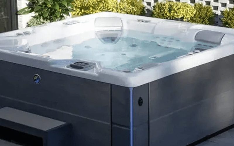 A modern outdoor hot tub with bubbling water sits on a stone patio next to a building, surrounded by plants and a decorative tiled wall. Steps lead up to the spa for easy access.