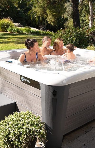 Four people relax and smile in a modern outdoor hot tub surrounded by greenery, trees, and stone paving on a sunny day. Steps lead up to the tub, and a potted plant sits nearby.
