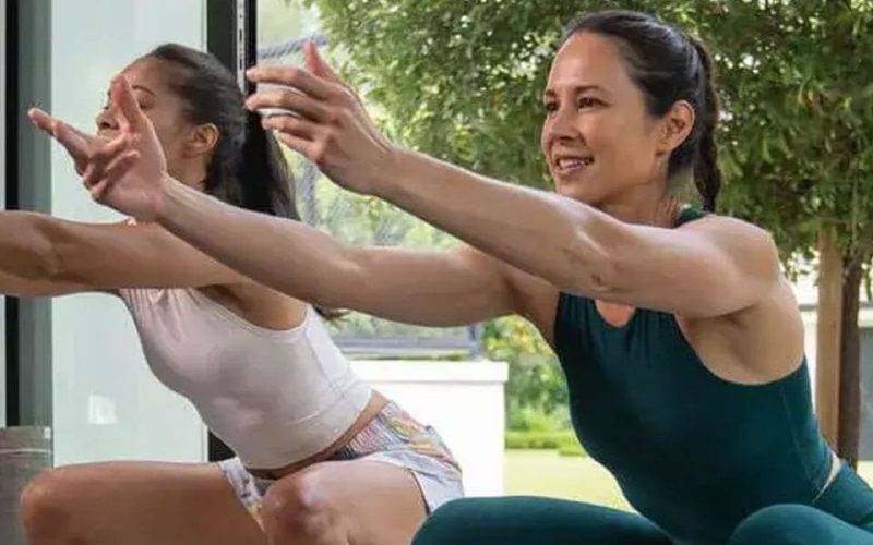 Two women in athletic wear are doing squats indoors near a large window, smiling and reaching forward with their arms extended. A tree and greenery are visible outside.