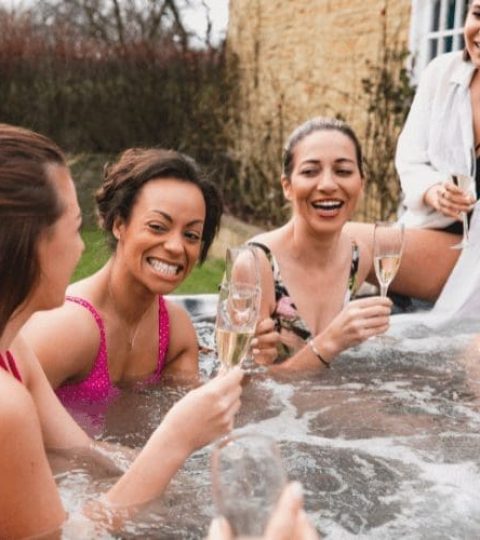 Four women in swimsuits sit together in a hot tub outdoors, smiling and holding glasses of champagne, enjoying a fun and relaxed moment.