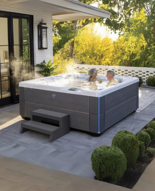 Two people relax in a steaming hot tub on a sunny patio beside a modern house, surrounded by greenery, with steps leading up to the hot tub and glass doors in the background.