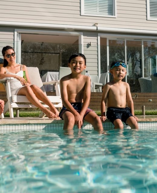 A family of four relaxes by a backyard pool. Two children sit on the pool’s edge with their feet in the water, while two adults lounge on chairs nearby in front of a modern house on a sunny day.
