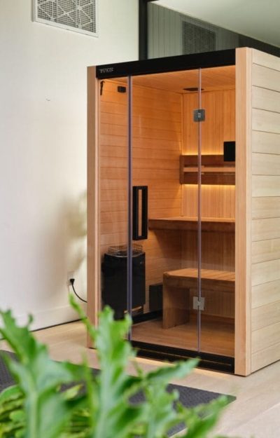 A modern, light wood sauna with glass doors is set indoors next to a large window, allowing views of lush green plants and the outdoor garden. The sauna features interior lighting and a wooden bench.