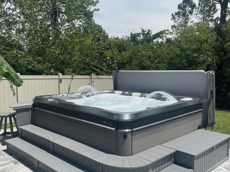 A gray hot tub with bubbling water sits on a raised deck with steps, surrounded by a fenced backyard with trees and greenery. A small orange object is on the steps, and the sky is partly cloudy.